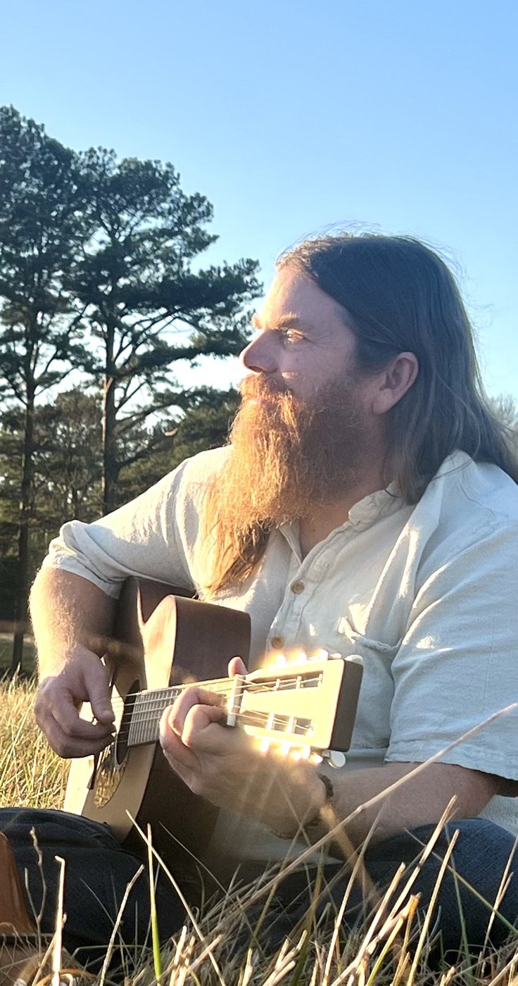 Drew playing acoustic guitar in the grass under the pines