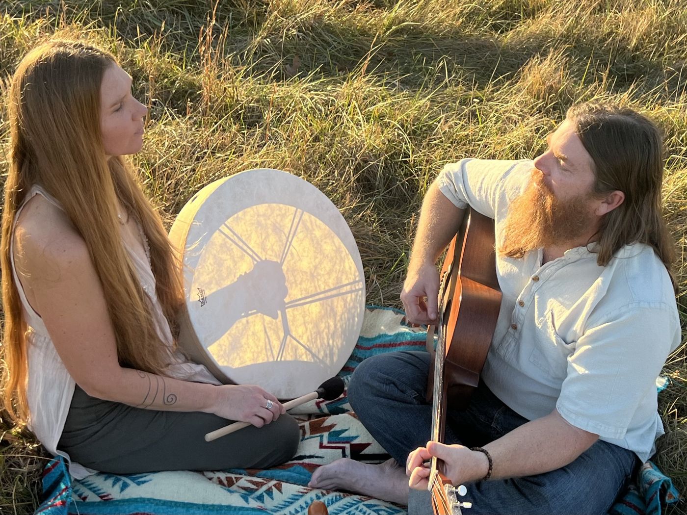 Destiny with a frame drum and Drew with an acoustic guitar, sitting on a blanket in tall grass