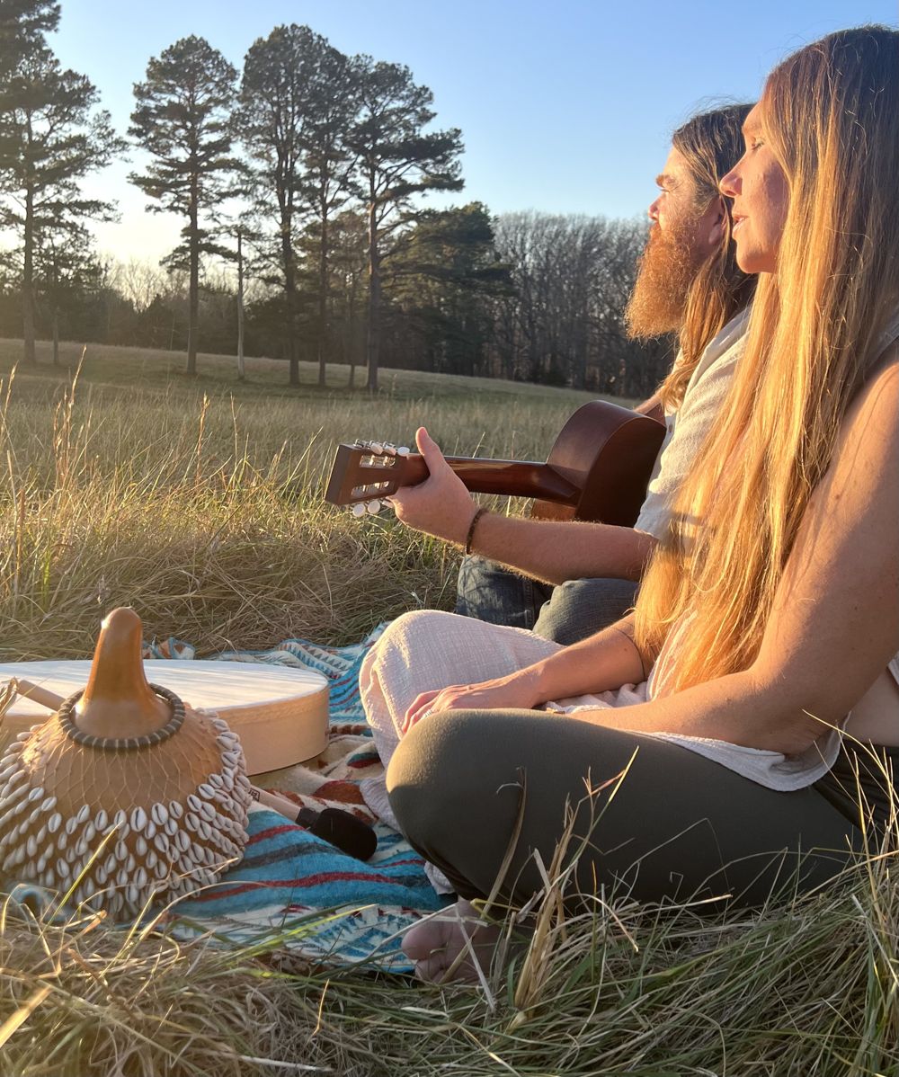 Destiny and Drew playing guitar and hand drum in a meadow at golden hour